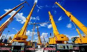 A row of yellow telescopic mobile cranes with booms raised against a blue sky