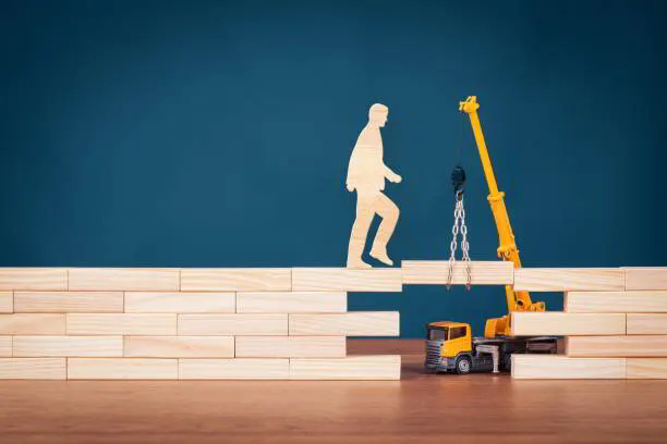 Wooden figure climbing a row of blocks next to a toy mobile crane, representing step-by-step progress