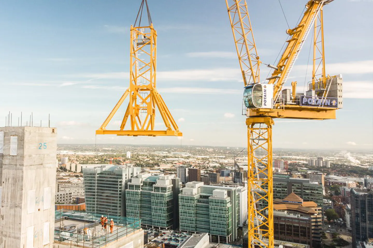 A tower crane lifting a steel section above a concrete high-rise core with a city in the background