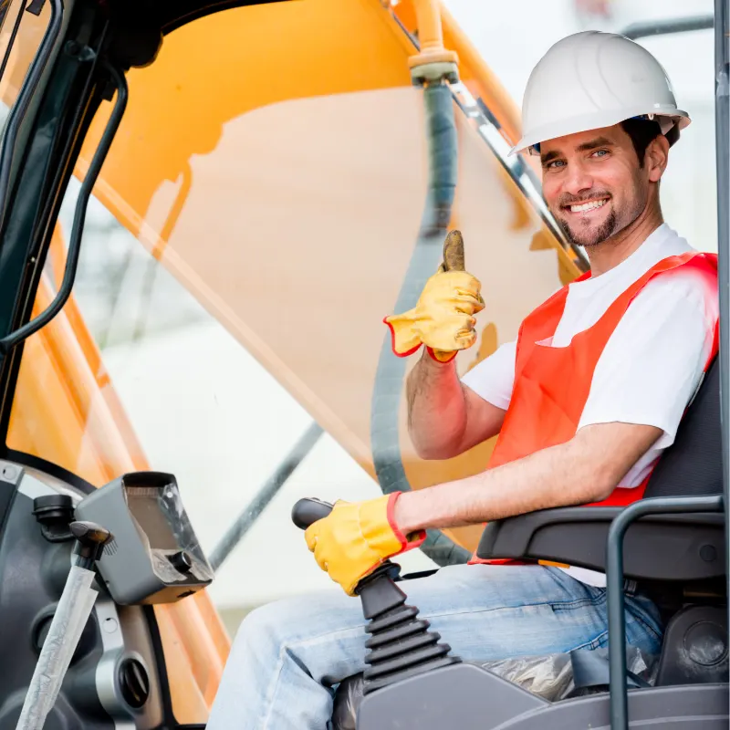 Smiling equipment operator in a white hard hat giving a thumbs up from the cab