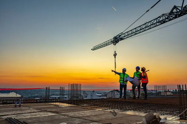 Three workers in hard hats reviewing plans on a rooftop under a tower crane at sunset