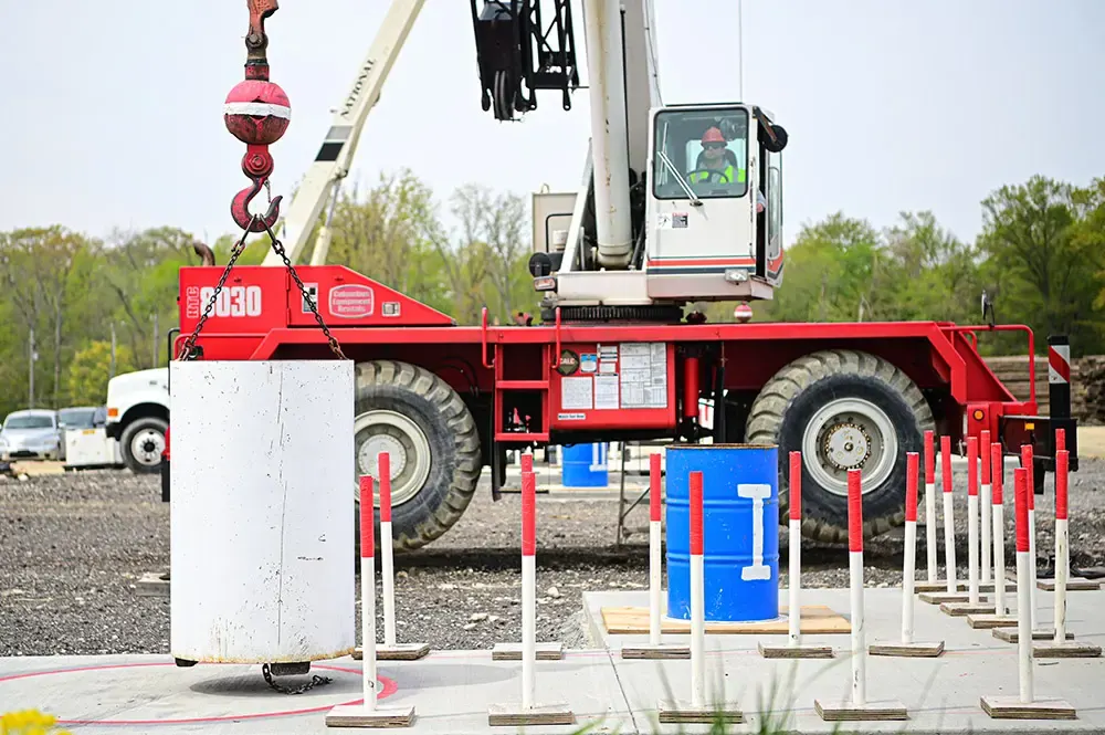 A rough-terrain crane on a practical exam course with a test weight hanging near red and white poles