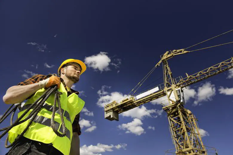 Worker in a hard hat and high-visibility vest carrying wire rope slings below a tower crane
