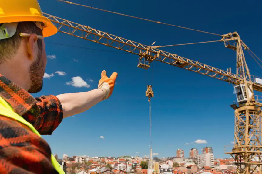 Worker in a hard hat giving a hand signal to a tower crane operator above a city skyline