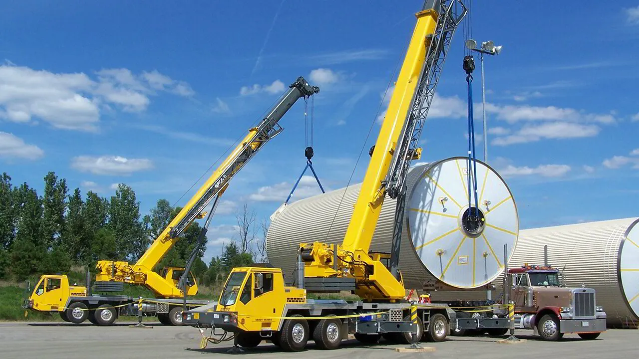 Two yellow telescopic truck cranes lifting a large cylindrical tank section