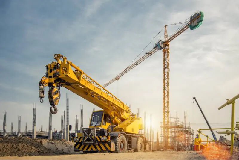Yellow rough-terrain mobile crane in the foreground with a tower crane rising behind it on a construction site